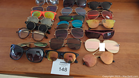 Top-down view showing 20 sunglasses arranged in rows on a wooden surface, showing variety of frame styles and lens colors, some with tags