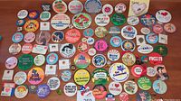 Wide view of about 50 colorful vintage buttons and pins spread out on a wooden surface showcasing text slogans and graphics.