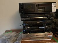 Stacked view of Yamaha cassette deck on top, Mitsubishi VHS player below, two JVC VCRs beneath it and a silver VCR at bottom with a bag of remotes at left.