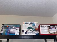Photo showing all three items in their original boxes: Sumo handheld vacuum on left, Black & Decker tub and tile scrubber in center, Shark handheld steam scrubber on right.