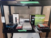 Wide view of lot items placed on black shelving unit showing various boxed and unpackaged electronics including a black wireless speaker base, Sony Android TV soundbar, Canary speaker, and data enclosures.