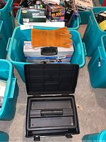 Two turquoise bins on concrete floor filled with tools; black case open in front, orange leather gloves and Mastercraft kit box visible