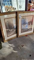 Two framed equestrian art prints with red and gold ornate wood frames leaning against a wall outdoors.