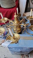 Brass six-light chandelier laying on a box showing arms, bulb sockets with faux candle sleeves, central body, and hanging chain. Plates and other items in background.