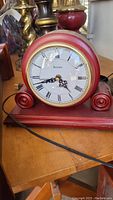 Front view of Bulova battery operated mantle clock with round clock face, Roman numerals, black hands, set in a red wooden frame resting on a wooden table.