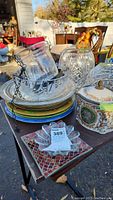Photo showing pile of plates, glassware, covered trinket box, and hanging candle holder on table outdoors.