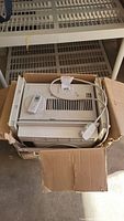 Top view of GE window air conditioner unit inside a cardboard box, showing power cord and remote control on top.