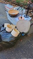 Five beige chandelier lampshades, a round wooden bowl with ball feet, KitchenAid slicer/shredder box, green frying pan, wine caddy, paper towel holders, placemats, tote bag and a large bag of faux greenery all set on a driveway surface