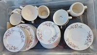 Overhead view of dinnerware stacked and organized in a bin showing plates, cups, saucers, and serving pieces with floral leaf pattern.