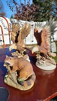 Three ceramic figurines: Lefton eagle on branch, Homco eagle with repaired feather, and Homco raccoon figurine on table outdoors.