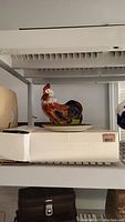 Colorful ceramic rooster figurine on a shelf with white box beneath