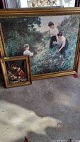 Two framed oil paintings leaning against a wall. Larger painting depicts three children in a dense field; smaller painting depicts Madonna and Child religious scene in a gold frame.