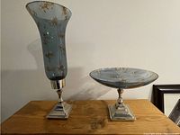 Front view of a tall decorative blue glass vase with gold detailing and a matching shallow decorative bowl, both on silver-tone square pedestal bases. Positioned on a wooden surface against a white wall.