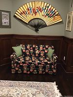 Front view of two-seat sofa with botanical pattern, two green throw pillows, in room with wood paneling and Asian style fan on wall
