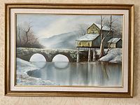 Framed oil painting showing a winter river scene with stone bridge and house, signed at the bottom left corner.