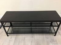 Front view of black metal TV stand with two glass shelves on light wood floor