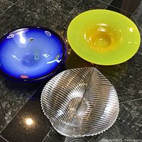 Three fruit platters on a dark granite surface seen from above: yellow glass, blue glass with colorful embedded designs, and clear ribbed glass.