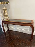 Straight-on view of rosewood console table with glass top and decorative lamp on left corner.