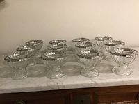 Twelve clear glass shrimp cocktail glasses with silverplate rims arranged in rows on a marble surface.