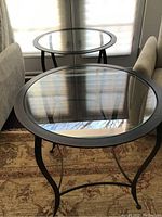 Two round tables made by Charleston Forge with black metal frames and clear glass tops, shown together on a rug between two sofas.