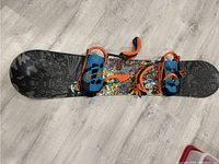 Snowboard featuring black and colorful graphics with blue and orange bindings attached, showing condition and features.