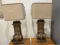 Pair of two wooden table lamps on wooden surface with beige rectangular fabric shades and rustic limed gracewood carved bases on clear acrylic feet.
