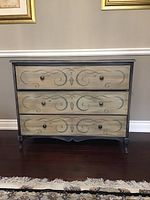 Front view showing entire chest with three drawers and scroll motifs on a dark wood floor.