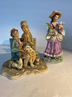 Two hand-painted porcelain figurines against a blue background: seated elderly couple and standing young woman