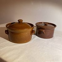 Two brown stoneware hand-thrown casseroles with lids, one larger than the other, shown side by side on white surface