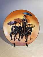 Front view of the Beatles ‘65 limited edition porcelain plate showing the four Beatles holding umbrellas against an orange background, sitting and standing on chairs, with a gold rim.