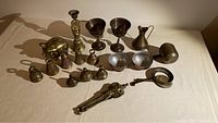 Overview of all items in the lot on a beige cloth background showing the full assortment including bells, goblets, utensils, piggy bank, door knocker, and paperweight