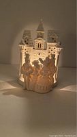 Front view of white bisque porcelain candle holder with a group of carollers in front of a castle showing the light glowing through.