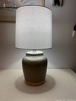 Full view of the ceramic table lamp on a flat surface with the white fabric shade illuminated showing the height and general shape of the lamp.