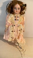 Full view of porcelain doll Gisselle in detailed pink satin dress with lace and ribbons, showing painted face and brown curly hair.