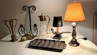 Full view of all six items arranged on a table: black metal candle holder, cast iron cornbread mould, brass dolphin tray, two mini lamps, and the claret pitcher
