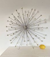 Front view showing the full starburst metal wall sculpture with glass orb ends, set against a white wall with a lemon placed beneath for scale.