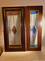Two stained glass cabinet doors standing side by side. Left door with rose central diamond glass, right door with blue diamond glass and wooden frames.