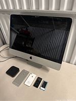 Apple iMac with screen off, two iPhones, and two iPods on table for parts or repair