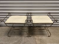 Pair of metal benches with curved legs and scroll detailing, each topped with a cream-colored cushion.
