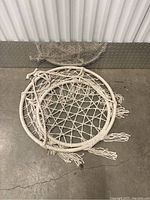 Photo of a white woven hammock chair with a circular frame and fringe tassels laid flat on concrete.