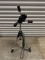 Full view of silver aluminum Manfrotto tripod with pan-and-tilt fluid head, quick-release leg locks, and carrying strap.