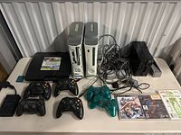 Photo showing two white Xbox 360 consoles, black PlayStation 3 and 2 consoles, black Nintendo Wii console, multiple controllers, and visible game cases, with cables.