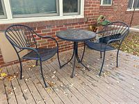 Full view of black metal bistro table with two matching chairs on wooden deck with autumn leaves scattered around.
