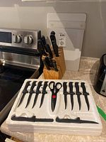 Wooden butcher block knife holder with knives and a set of 8 steak knives and scissors in white packaging on a kitchen counter.