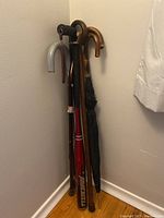 Photo showing three wooden canes, a black umbrella, and a red Worth aluminum baseball bat grouped together in a corner.