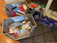 Two gray plastic storage bins containing various cleaning and household products including brushes, sprays, and soap bottles. Several blue bottles of Finish dishwasher cleaner are visible alongside dish soap and Vim.