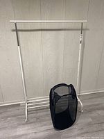 White metal clothes rack on casters with black folding mesh laundry hamper set inside, against a light wall.