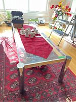 View of the chrome and glass coffee table in a bright room showing the glass top, chrome legs and frame, with a red table runner and some ceramic decor on top, with the chip on chrome frame visible on one corner.