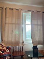 Two beige linen weave curtain panels hanging on a curtain rod in front of a window. Panels have a lightweight semi-sheer texture and metal grommets along the top edge.