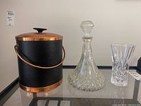 Three-piece set showing black ice bucket with copper accents on left, glass decanter with stopper center, and crystal-cut glass vase on right.